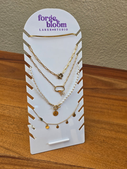 A stack of four delicate gold and silver chain necklaces with small charms, displayed on a white card stand branded 'forge & bloom LASER STUDIO' on a wooden table.
