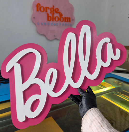 Hand holding a pink and white 'Bella' sign with 'forge bloom' in the background.