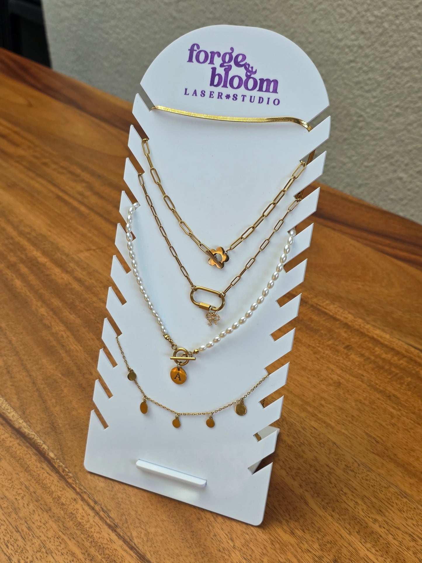 A three-layer gold necklace set with various charms displayed on a white card stand branded 'forge & bloom LASER•STUDIO'. The set includes a paperclip chain, a pearl bead chain, and a delicate chain with small gold teardrop charms, resting on a wooden table.