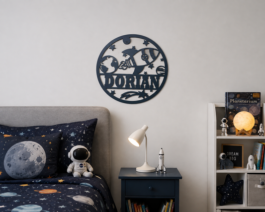 A circular dark navy metal wall sign with a laser-cut space theme and the name 'DORIAN' in the center, mounted on a light gray wall in a styled children's bedroom. The room features a bed with a dark navy space-print bedding set, a matching pillow, a small astronaut plush toy, a bedside table with a lamp, and a bookshelf with space-themed decor.