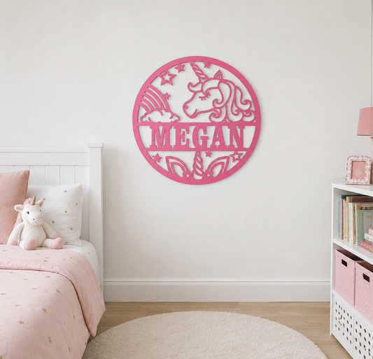 A pink circular wall art piece featuring a unicorn silhouette and the name 'MEGAN' in the center, mounted on a white wall in a styled children's bedroom with a bed, shelf, and rug.