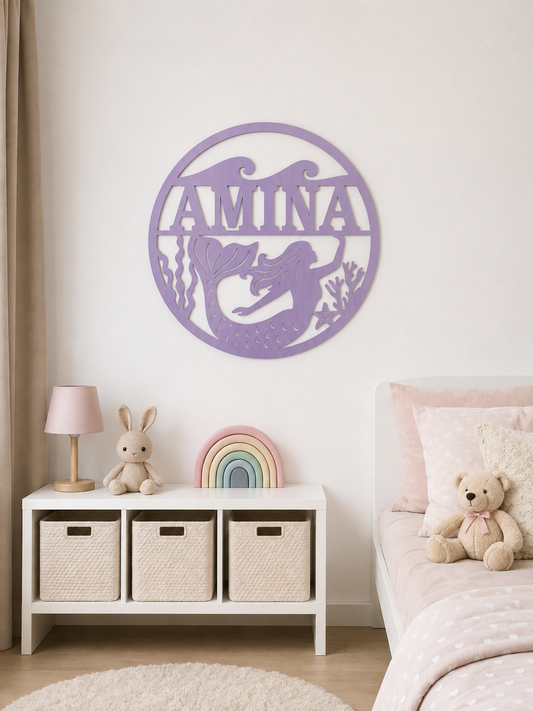 A purple circular wall art piece featuring a mermaid and the name 'AMINA' is displayed on a cream wall in a styled children's bedroom. Below the art, a white storage cabinet with three beige woven baskets holds a pink lamp, a plush bunny, a pastel rainbow, and a teddy bear, next to a bed with pink bedding.