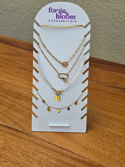 A three-layer gold and pearl necklace set with small charms, displayed on a white Forge & Bloom Laser Studio card, resting on a wooden table.
