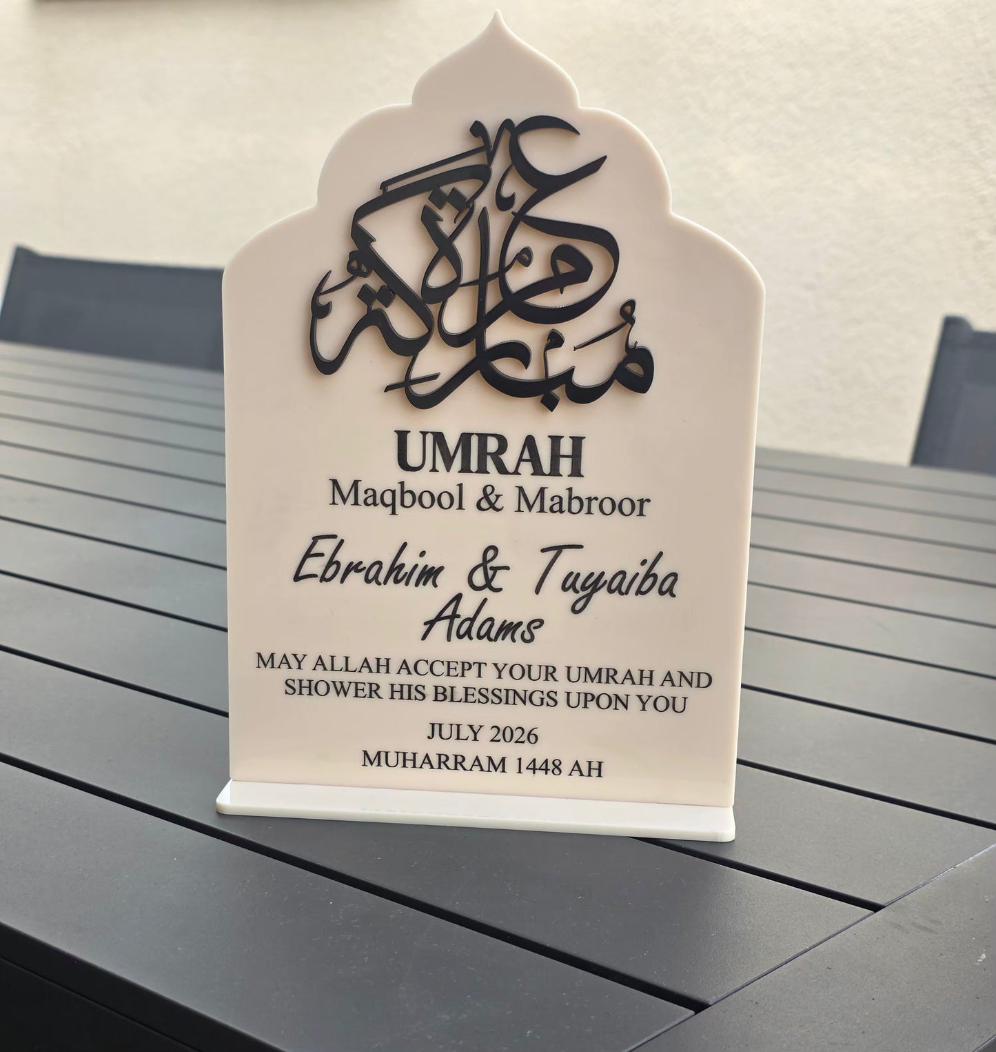 A personalized Islamic umrah plaque with black Arabic calligraphy and text on a white arched stand, placed on a dark gray slatted table.