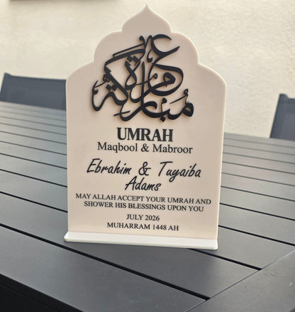 A personalized Islamic umrah plaque with black Arabic calligraphy and text on a white arched stand, placed on a dark gray slatted table.