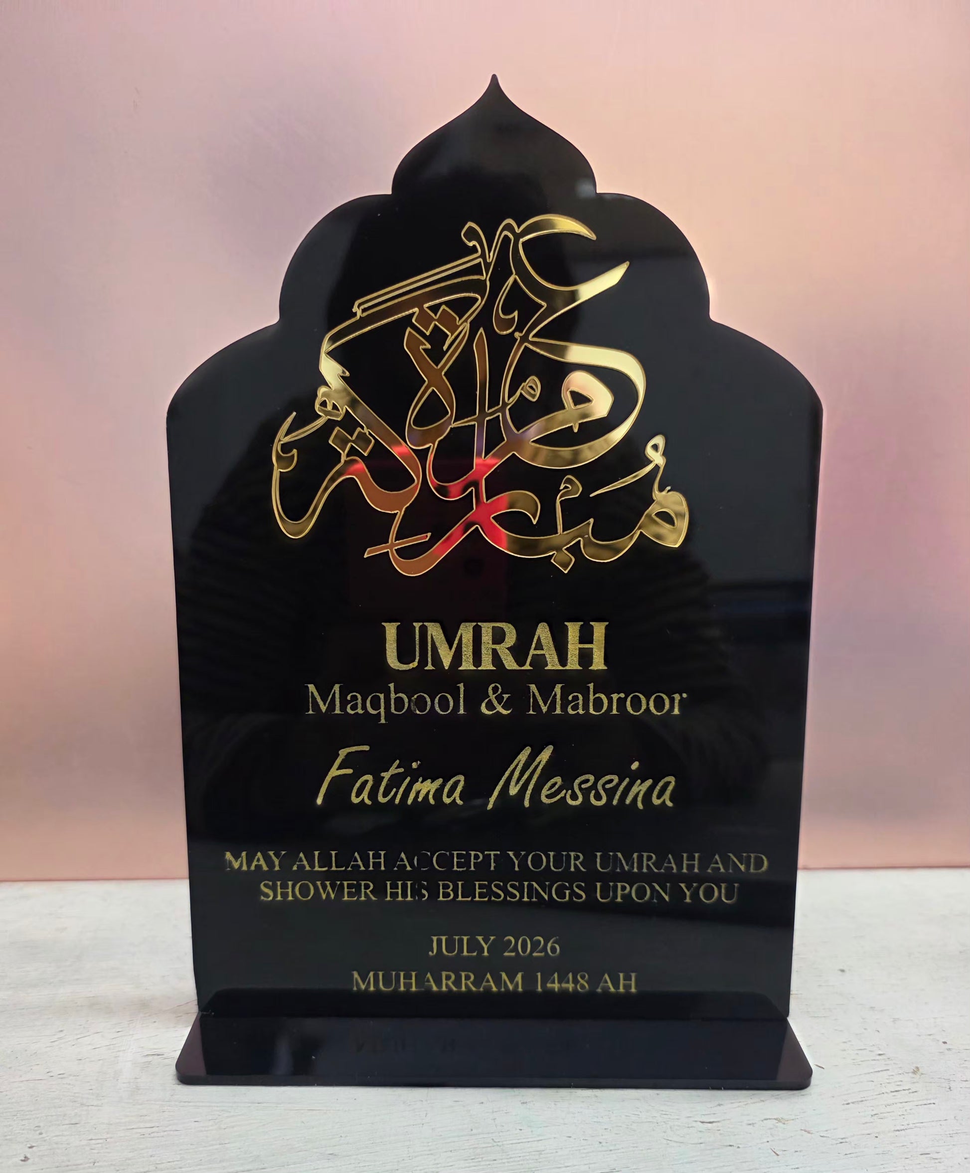 A black acrylic Islamic umrah plaque with gold Arabic calligraphy and personalized text, displayed on a light surface against a soft pink background.