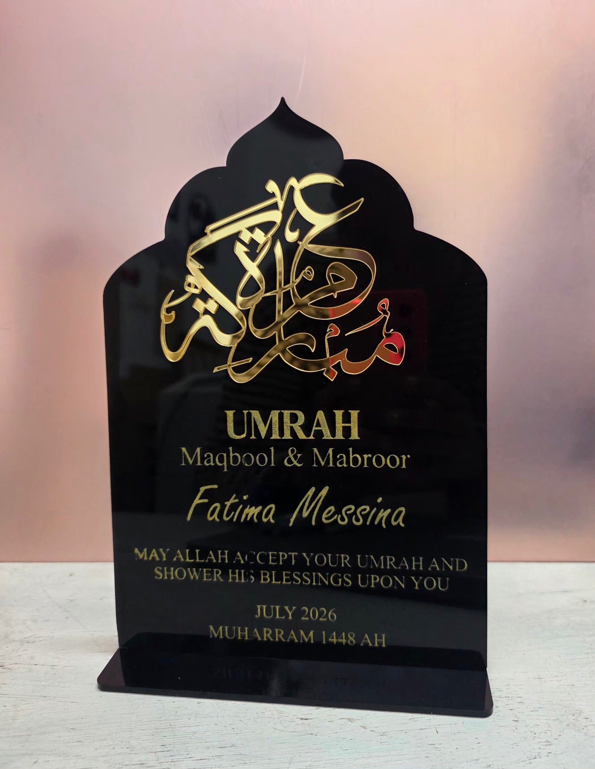A personalized black acrylic Islamic umrah plaque with gold Arabic calligraphy and custom text, displayed on a light surface against a soft pink background.